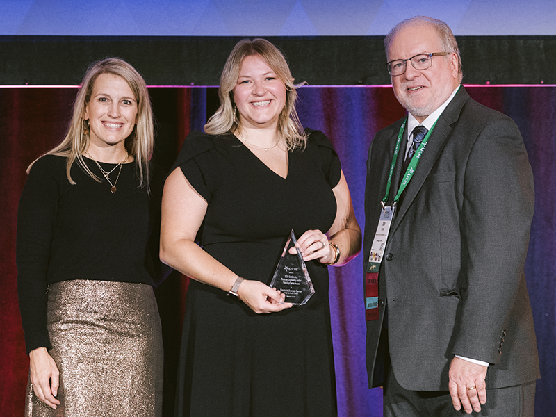 Three people stand on stage, with the central woman in a black dress holding a clear triangular award, flanked by a woman in a glittering skirt and a man in a suit with a conference badge.