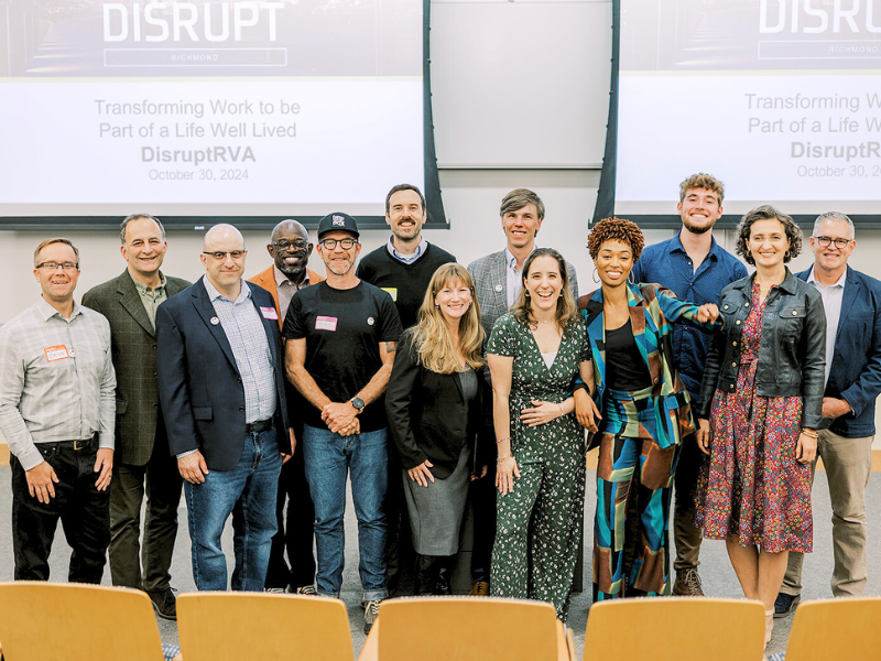 A group of professionals smiling and posing in front of a presentation screen at the "DisruptRVA" event on October 30, 2024, with the tagline "Transforming Work to be Part of a Life Well Lived."