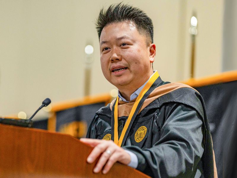 A speaker wearing a black academic gown and medal delivers a speech at a podium during a ceremony.