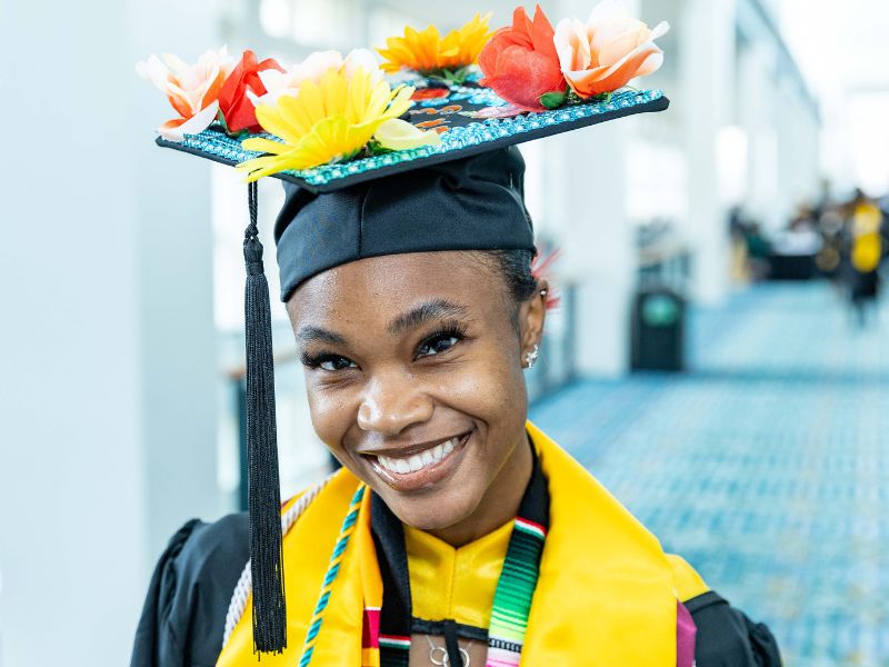 A smiling graduate wearing a decorated cap with colorful flowers and a yellow stole poses for a photo in a bright indoor venue.