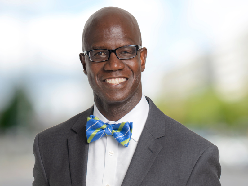 Portrait of a smiling man wearing glasses, a gray suit and a blue-and-green bow tie against a blurred background.