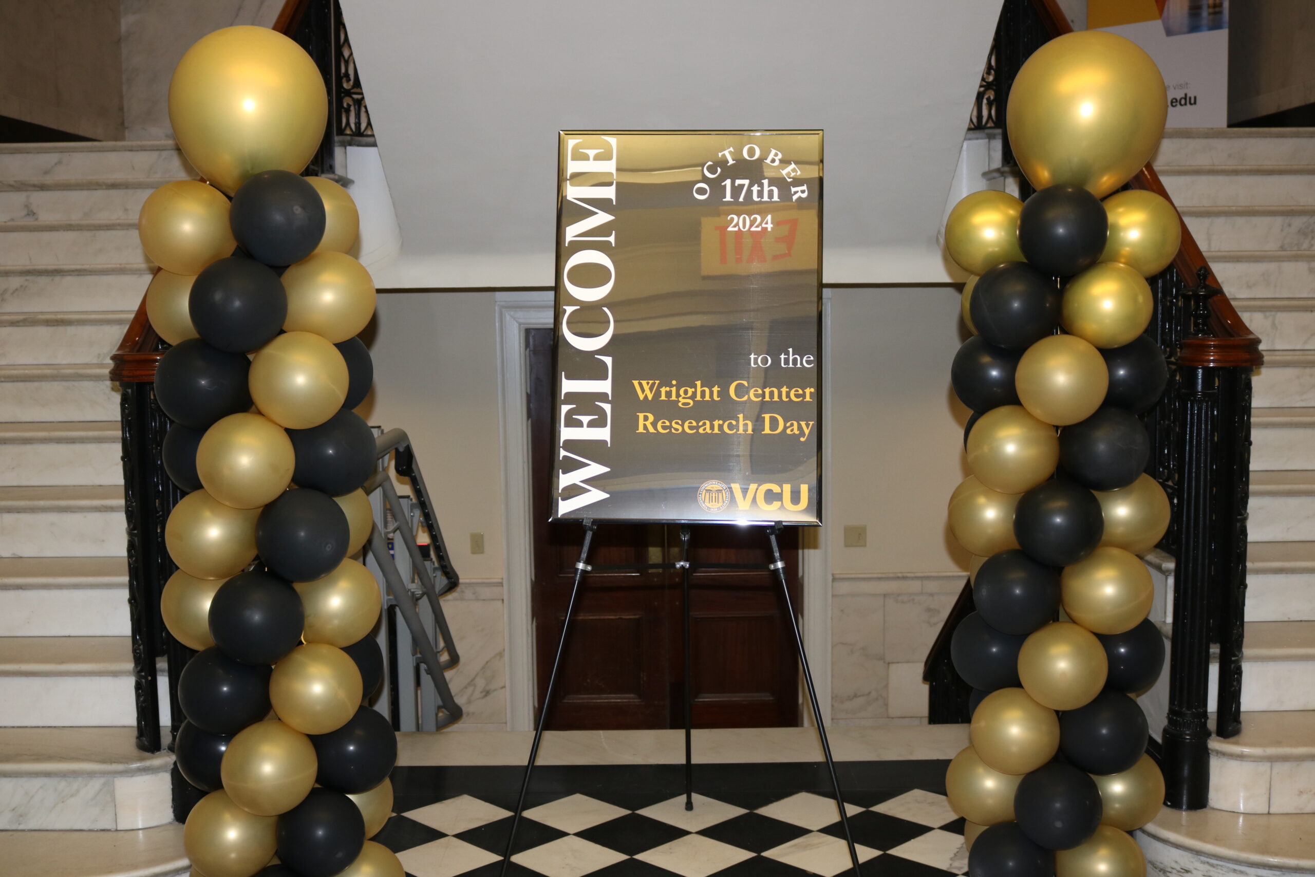 "A welcoming sign for the Wright Center Research Day on October 17th, 2024, displayed in a gold frame on an easel. The sign is surrounded by decorative columns of black and gold balloons, placed in a marble-floored entryway with a staircase in the background."