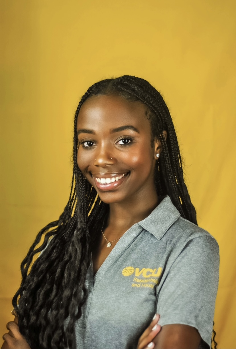 Victoria Chege smiles brightly against a bright yellow background. She wears a VCU Residential Life and Housing collared shirt.