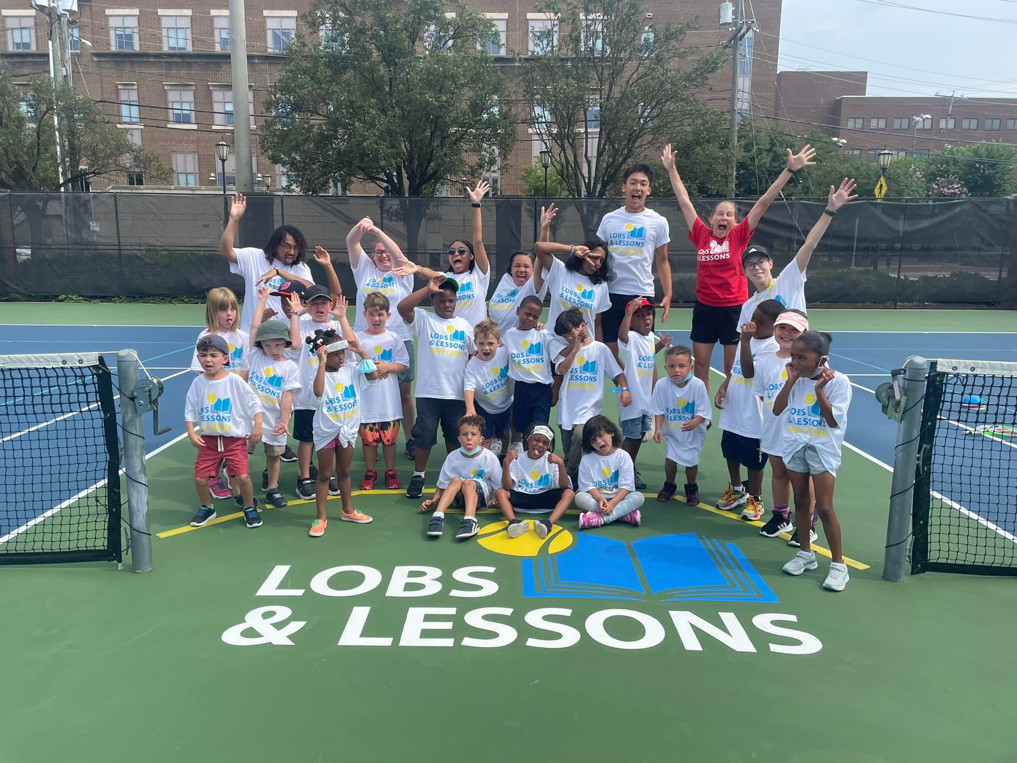 Lobs & Lessons staff on a tennis court with a large group of kids, all pulling silly faces and poses.