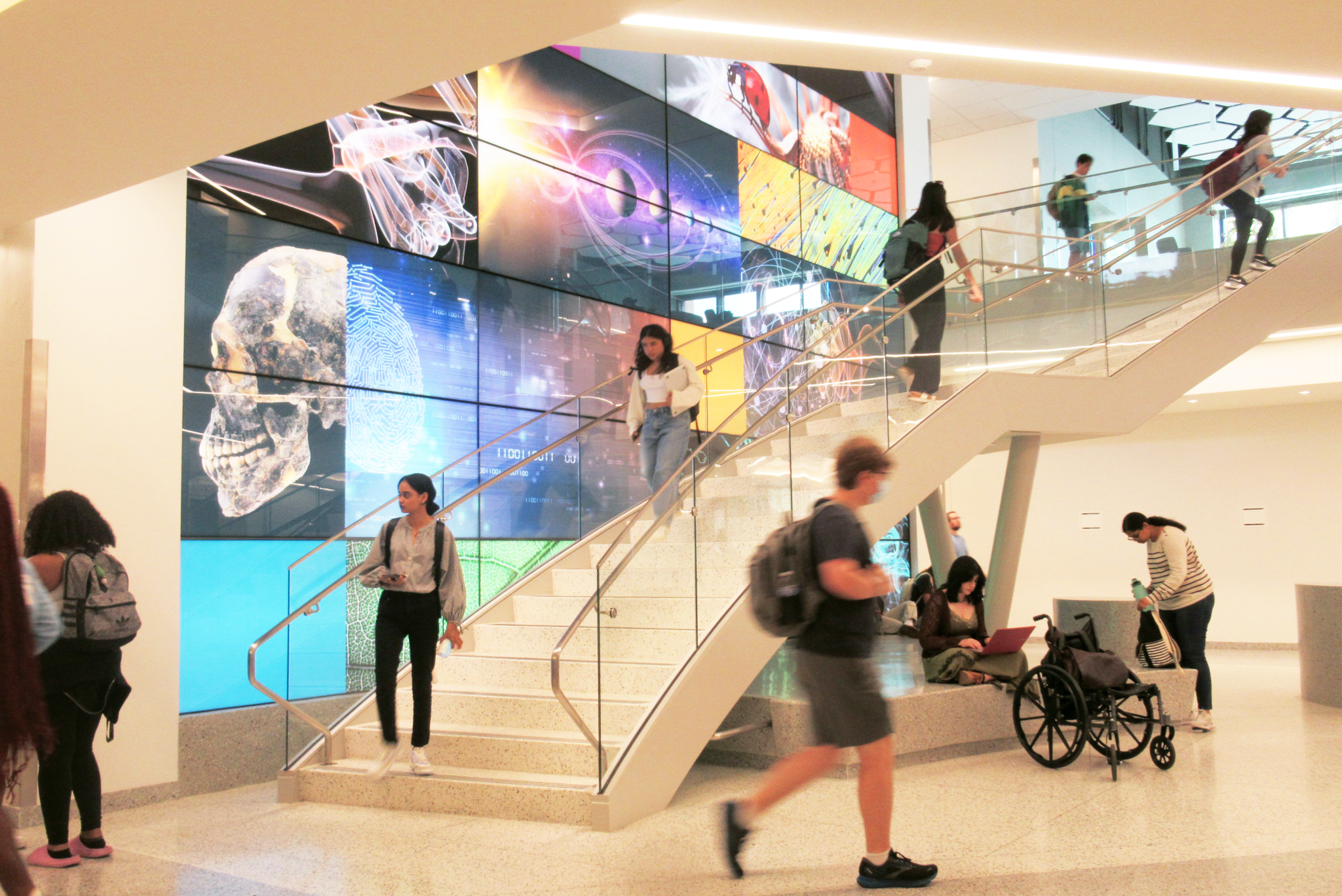 Students walk up and down stairs in an academic building. Up the wall are colorful screens with science-related imagery. Under the stairs, a student studies with their wheelchair besides them.