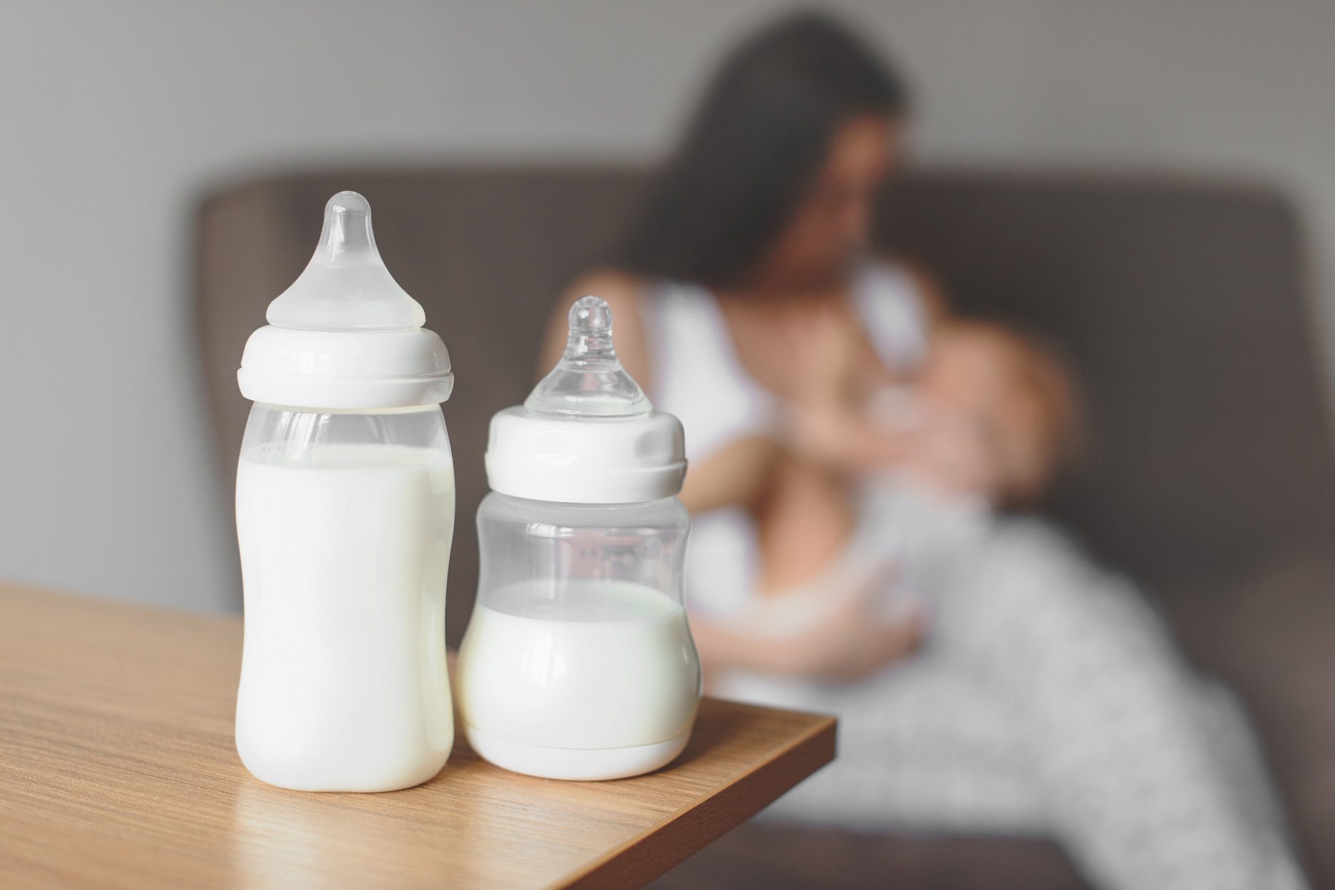 Two bottles of milk in the foreground. In the background is a blurry mother with a baby on her lap.