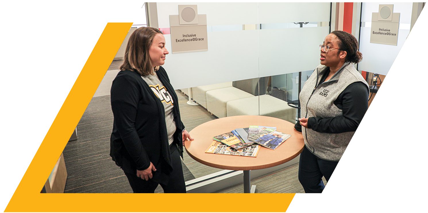 Two women stand in an office, speaking to each other around a circular table.