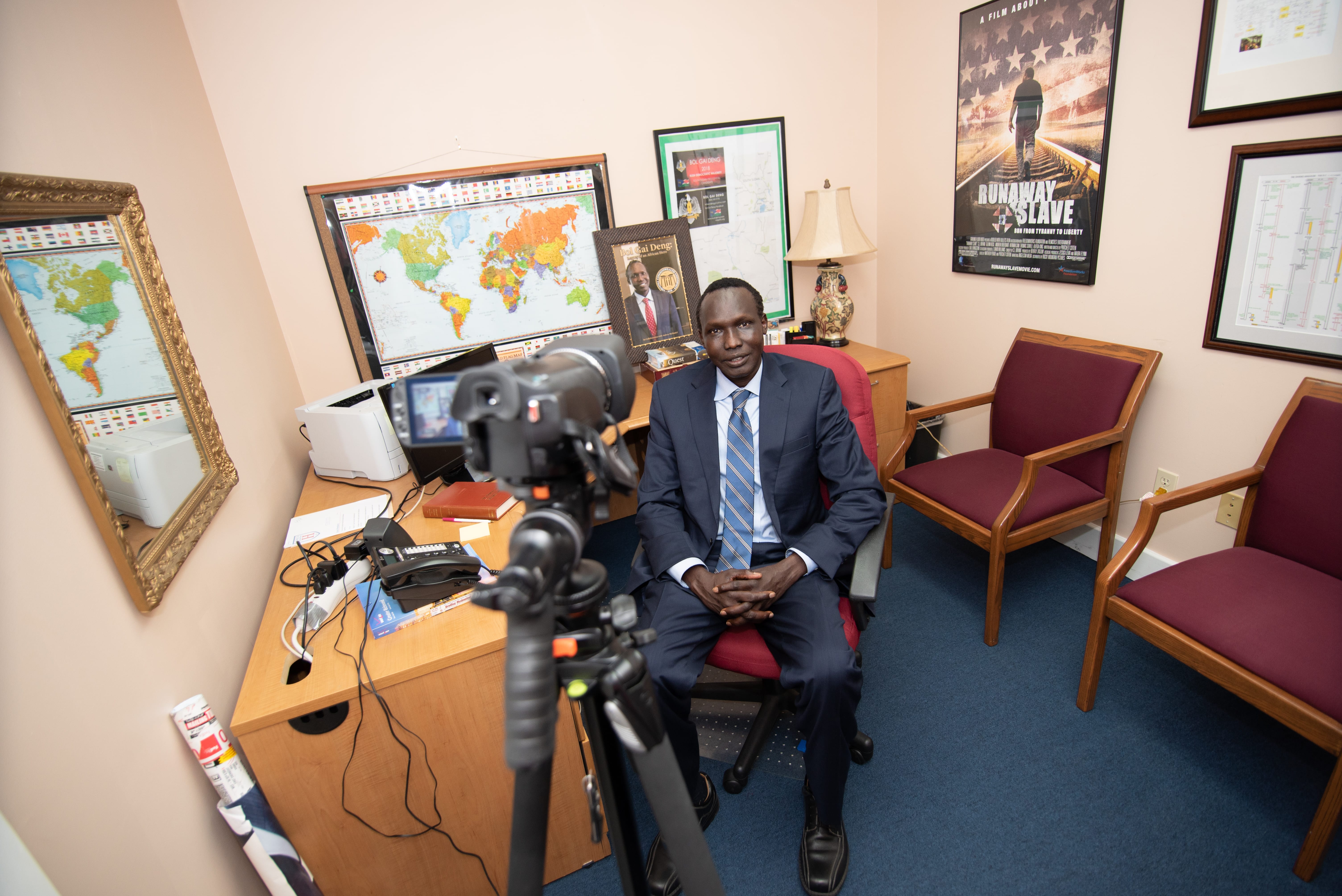 Bol Gai Deng sits in an office. In the background are world maps and a poster for the movie "Runaway Slave."