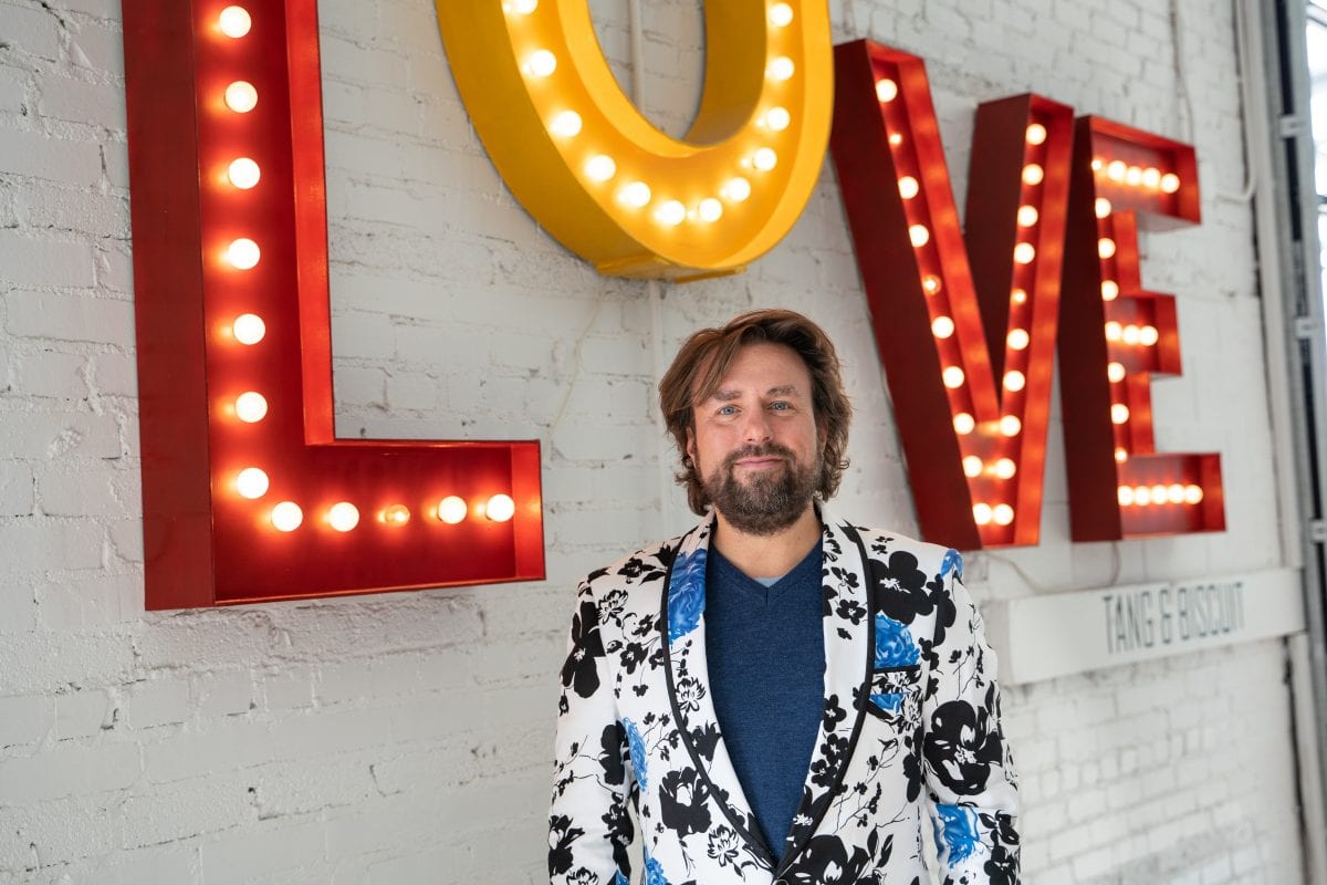 David Gallagher wears a colorful blazer as he stands beneath a large, lit sign proclaiming "love."