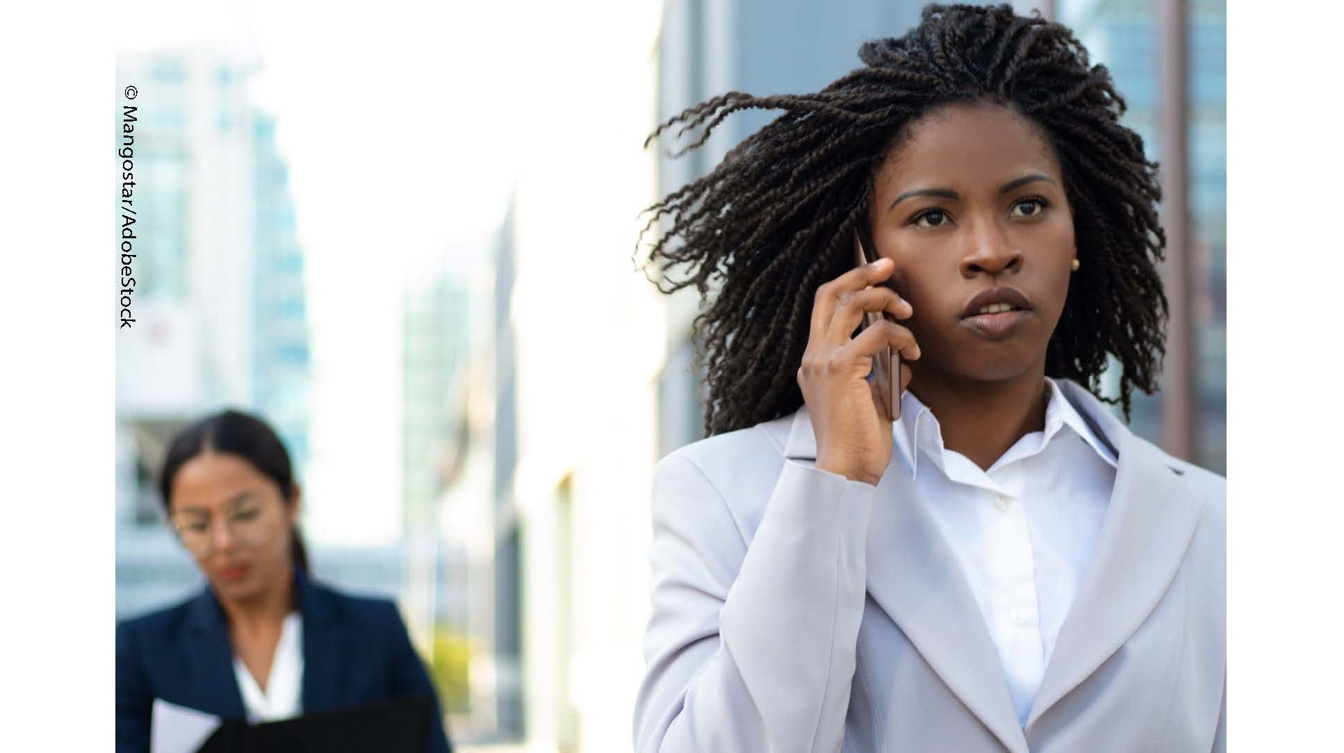 Ayana, a young woman in a business suit, is making a call on a cell phone. Another woman in glasses and a suit is standing in the distance, behind her.