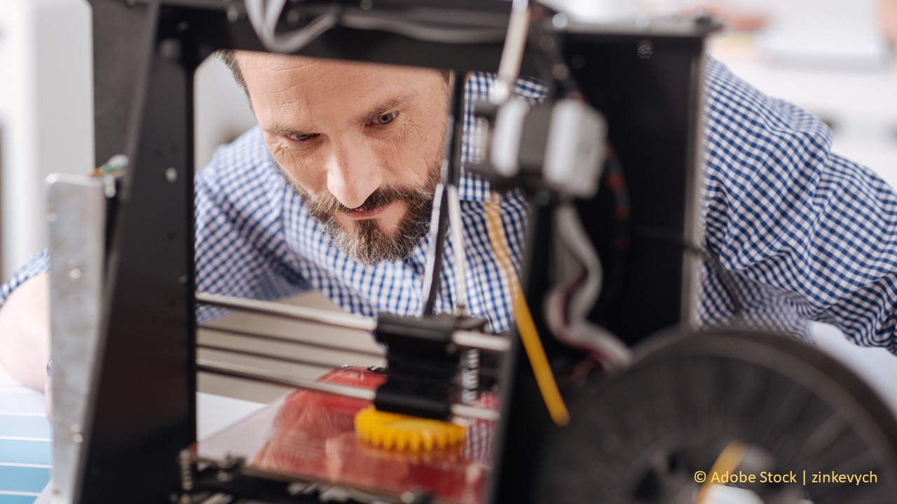 A man with brown hair and a beard is bending down to look into a 3D printer as it prints a small, yellow plastic item.