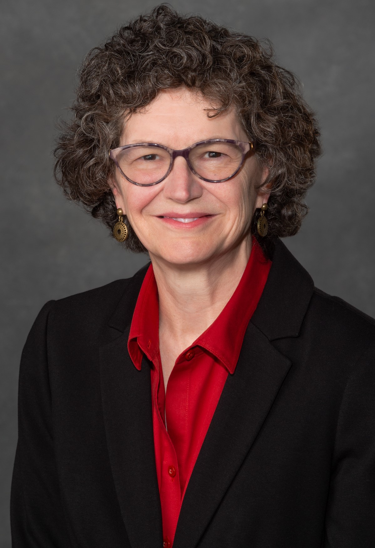 SUzanne Milton, Chief Ethics and Compliance Officer for VCU, smiles at the viewer. She has short, curly brown hair, glasses, and is wearing a bright red blouse and a black blazer with gold earrings.