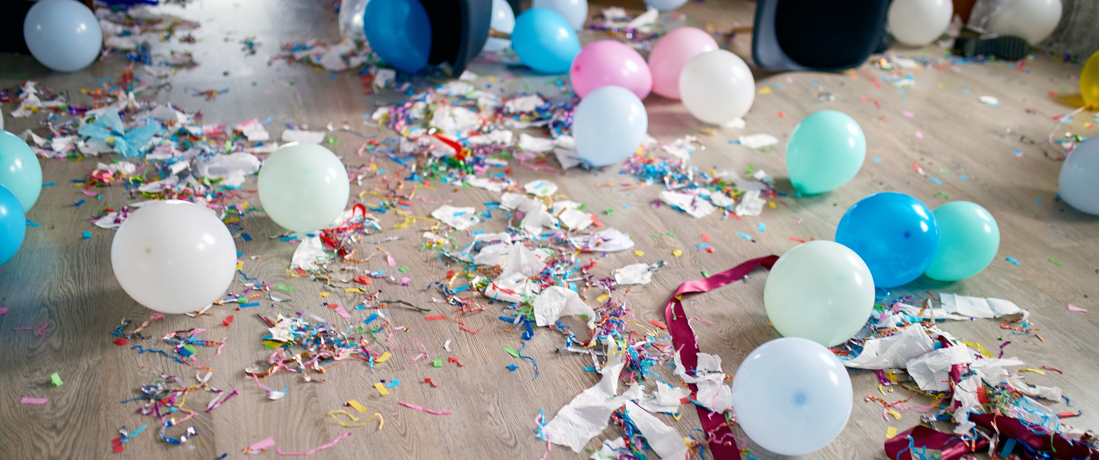 Trash from a party is scattered all over a wood floor. Balloons, streamers and confetti of different colors are everywhere.
