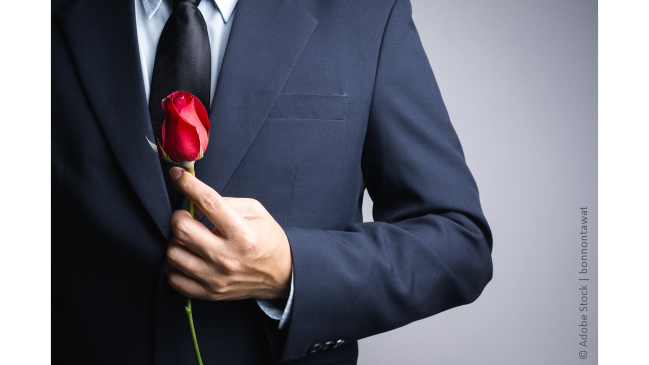 A man in a blue suit with a black tie holds a long-stem red rose close to himself, like he's about to surprise someone with it.