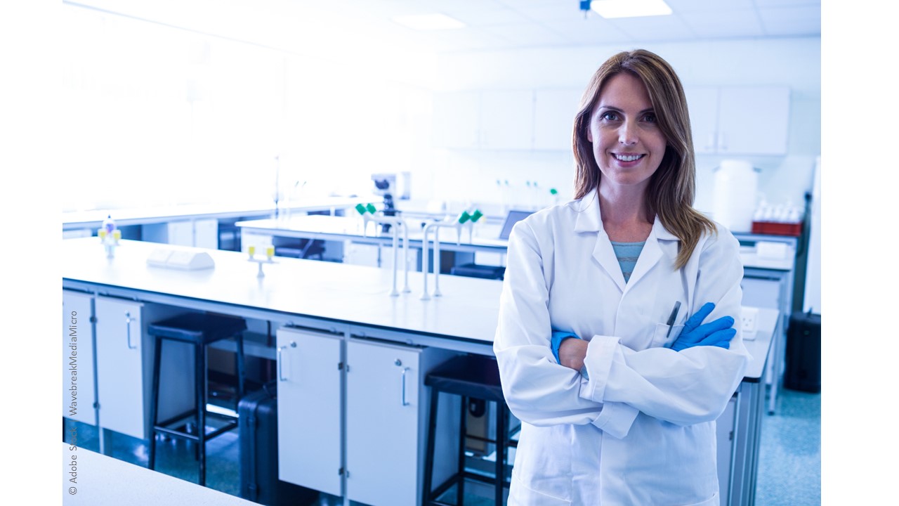 A woman who is a scientist and researcher stands in an empty laboratory, wearing a white lab coat and blue, disposable gloves. She is smiling at the viewer and her arms are crossed in front of her.