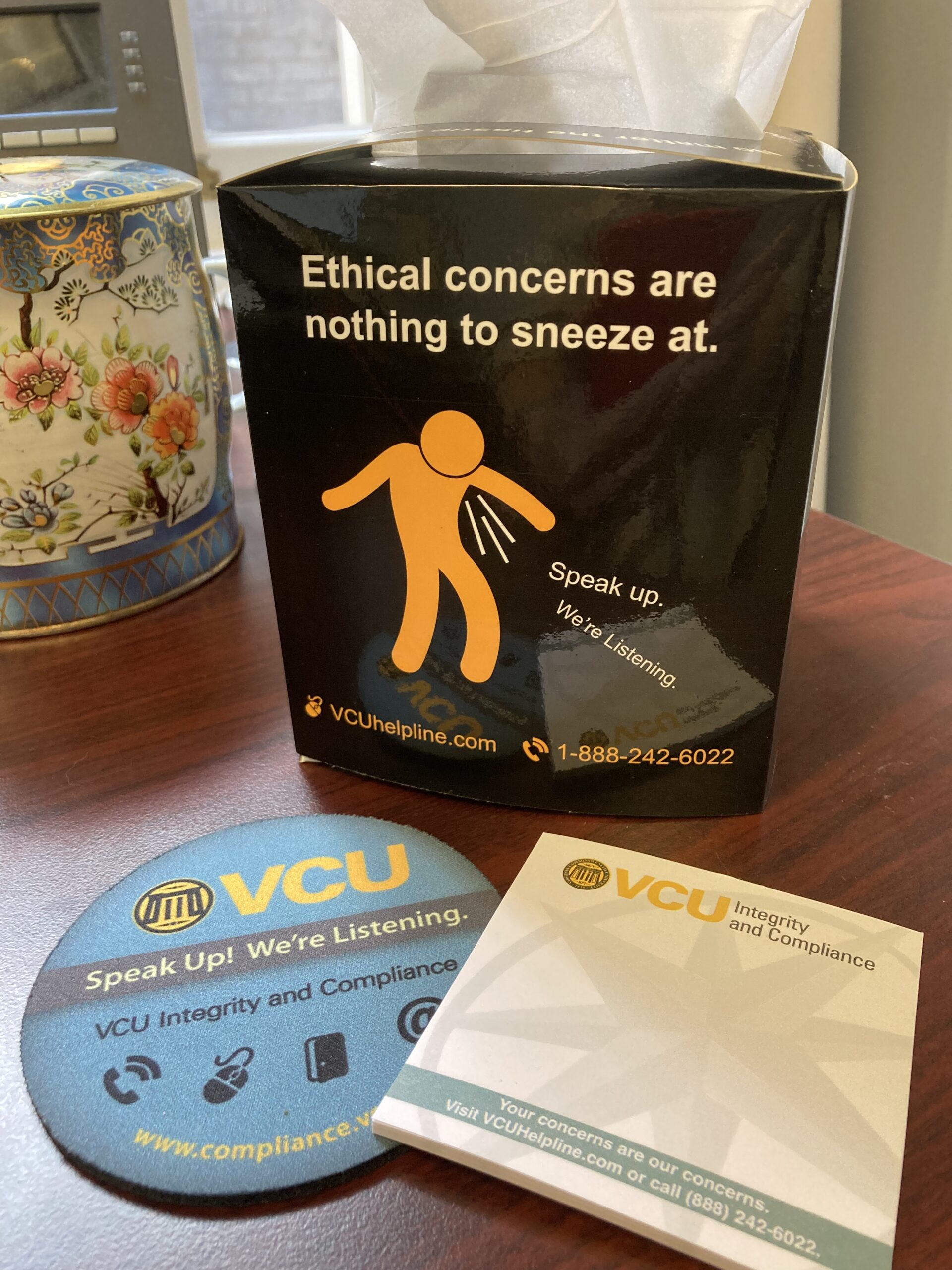 A desktop scattered with several tchotchkes from the Integrity and Compliance Office. A sticky-note pad says, "You're concerns are our concerns," a coaster says, "Speak up! We're listening," and a tissue box cover shows a picture of a stick figure that's sneezing, and says, "Ethical concerns are nothing to sneeze at."