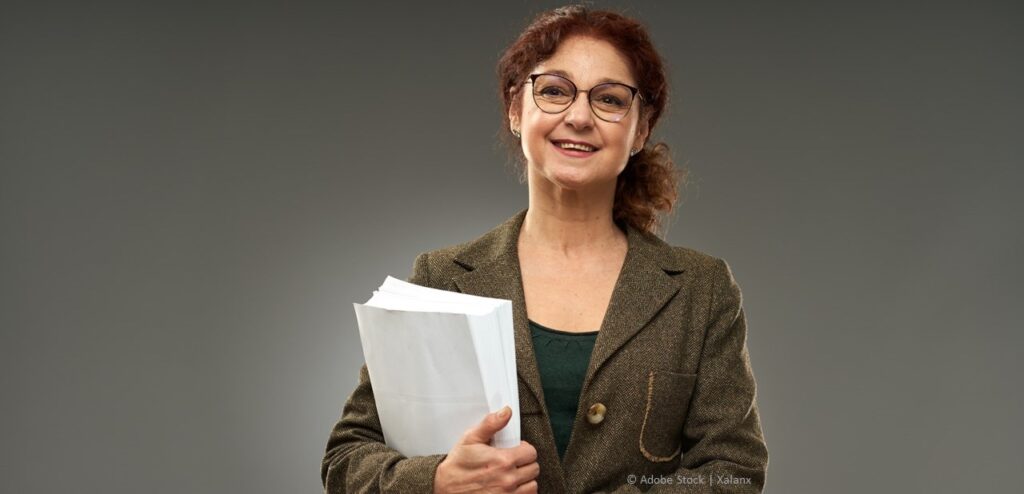 A middle-aged woman with glasses and red hair pulled back in a ponytail is holding a stack of papers and smiling.