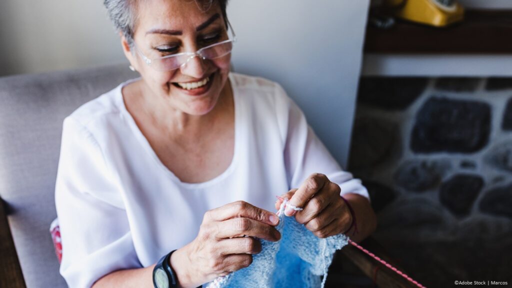A woman with short grey hair and glasses is smiling as she crochets a light blue baby blanket.