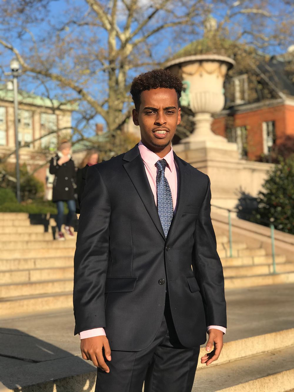 Matthews Tessema posing in a suit outdoors. Behind him are stairs, other people walking by, and buildings.