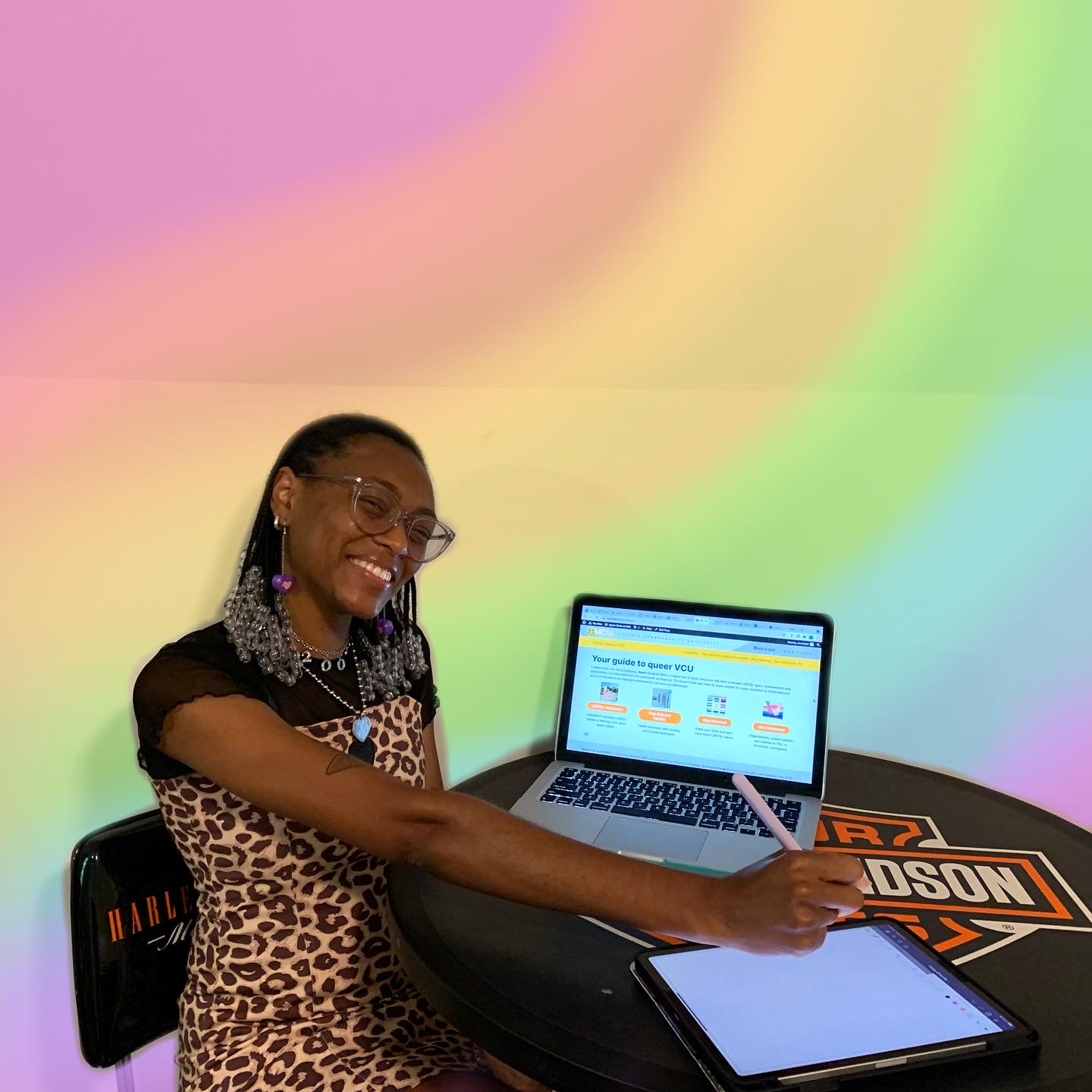 Murray smiles in front of a laptop screen displaying Queer Circle at VCU. The background has been edited with pastel rainbow colors.