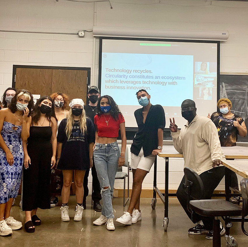 Guthrie's Fashion and Sustainability students pose in front of a screen with a presentation slide by community partner Circ. The students have masks on and some are holding up peace signs.