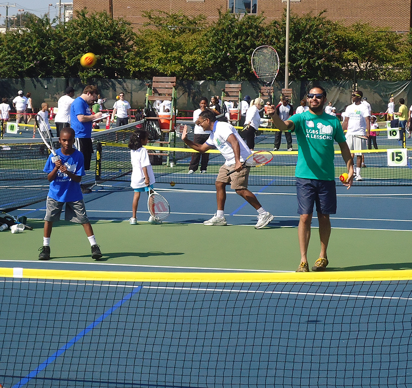 Families play tennis together outside at the Mary and Frances Youth Center tennis courts on a bright, sunny day.