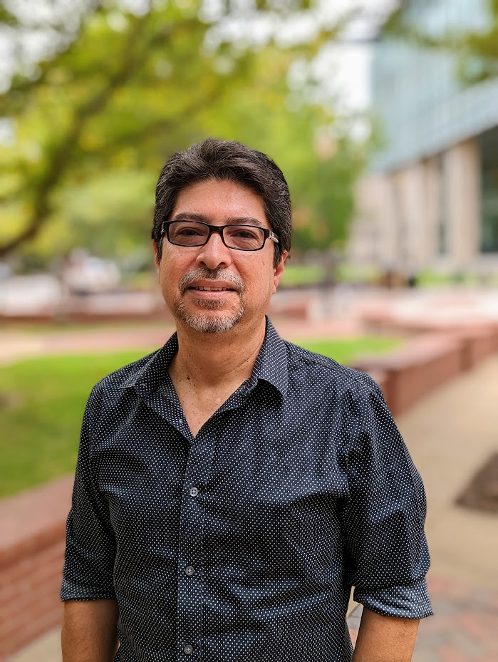 Carlos Escalante poses outside of the VCU Cabell Library. It's a bright, sunny day, and he wears glasses and a black button-up with a dotted pattern.