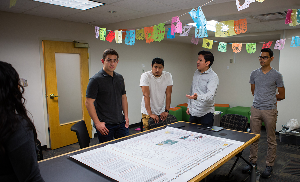 Moreno and three Latino students stand together in a room with colorful garland bunting strung on the ceiling. There is a table with a large poster showing VCU-affiliated research.