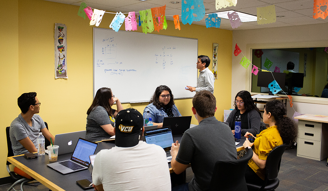 Dr. Moreno writes on a whiteboard in a room with half a dozen Latine students. Colorful, festive garland bunting is strung from the ceiling.