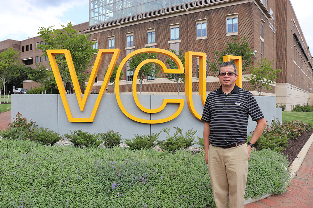 Fernando Tenjo-Fernandez stands outside in front of a large yellow VCU letters. There's green foliage and a VCU building behind him.