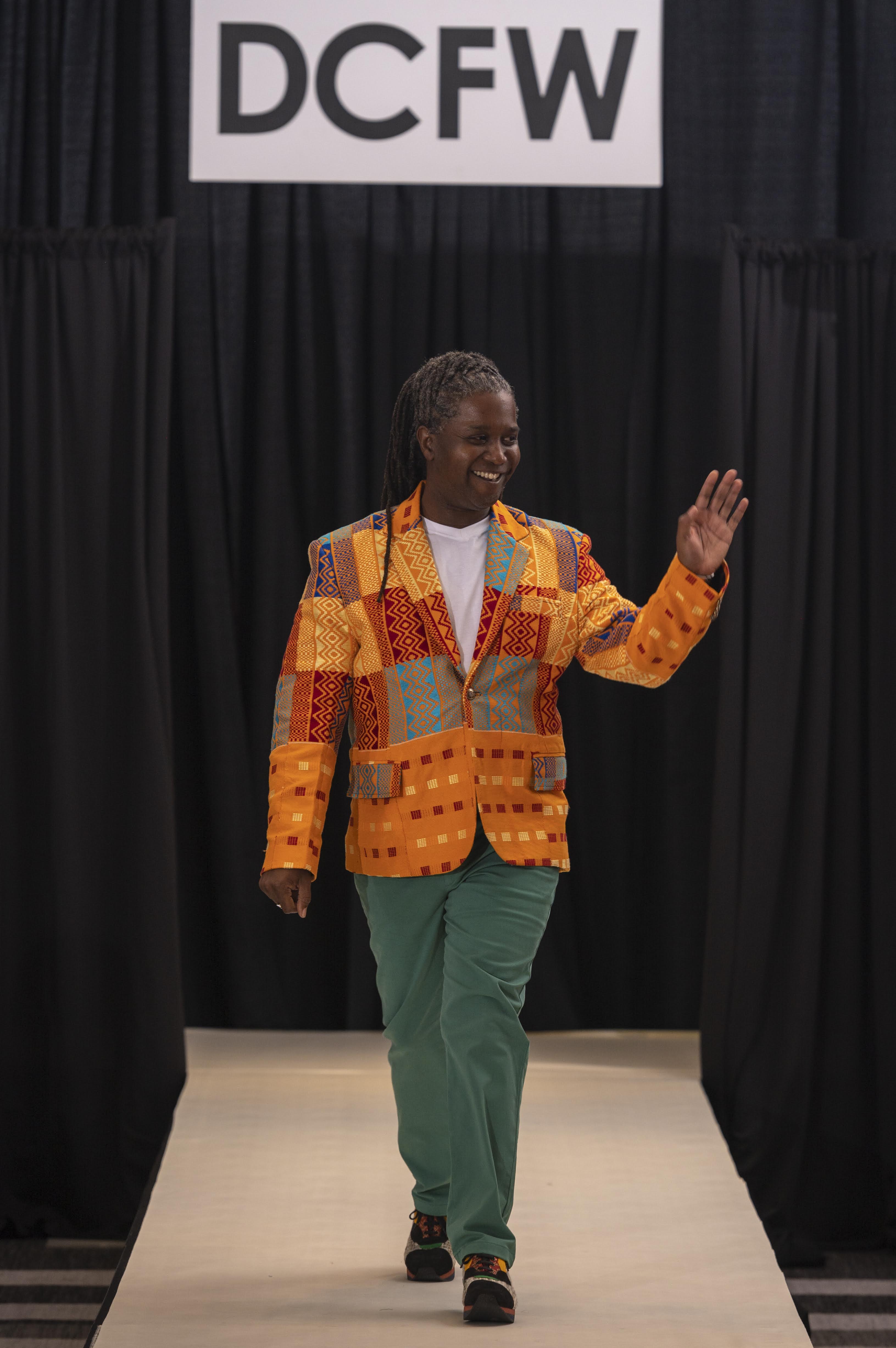 Gattis walks down a runway smiling and waving. He wears a bright orange patterned blazer with a white undershirt and green trousers. Behind him are black curtains and a large sign that says "DCFW."