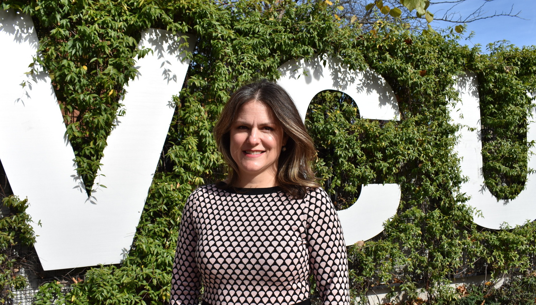 Dr. Lingold poses outside in front of a large white "VCU" sign with beautiful green foliage around it. She wears a printed long-sleeve dress.