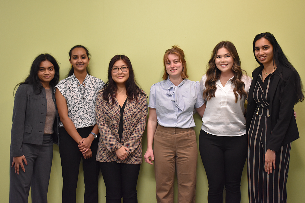 Six students pose together and smile against a blank yellow wall. They are all in professional dress clothes.