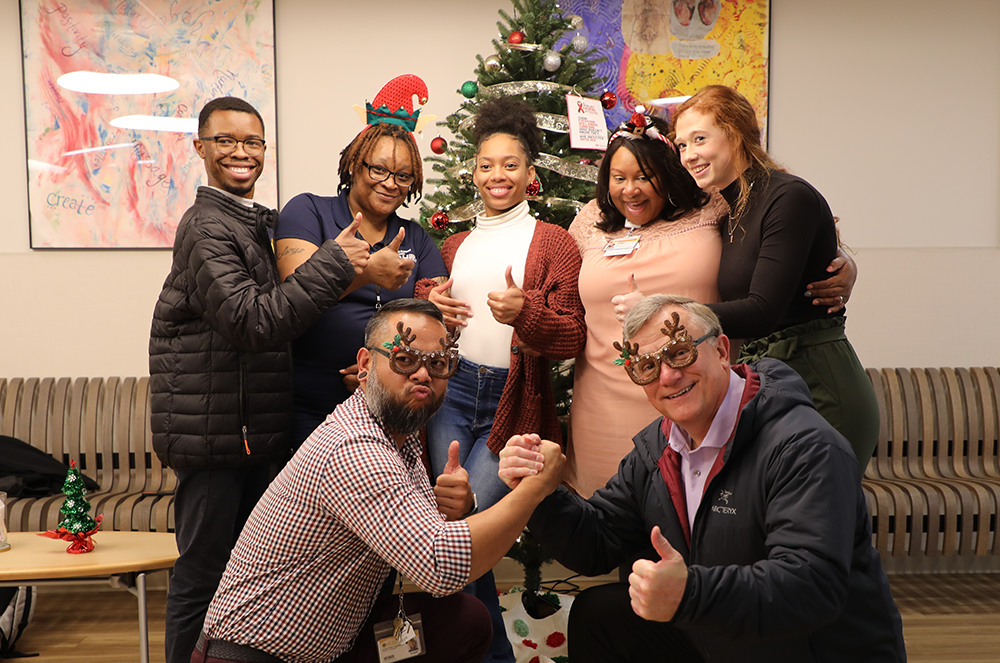 A group of VCU Health Hub staff and interns pose in front of a Christmas tree. Everyone is in various festive gear, from elf hats to reindeer sunglasses.