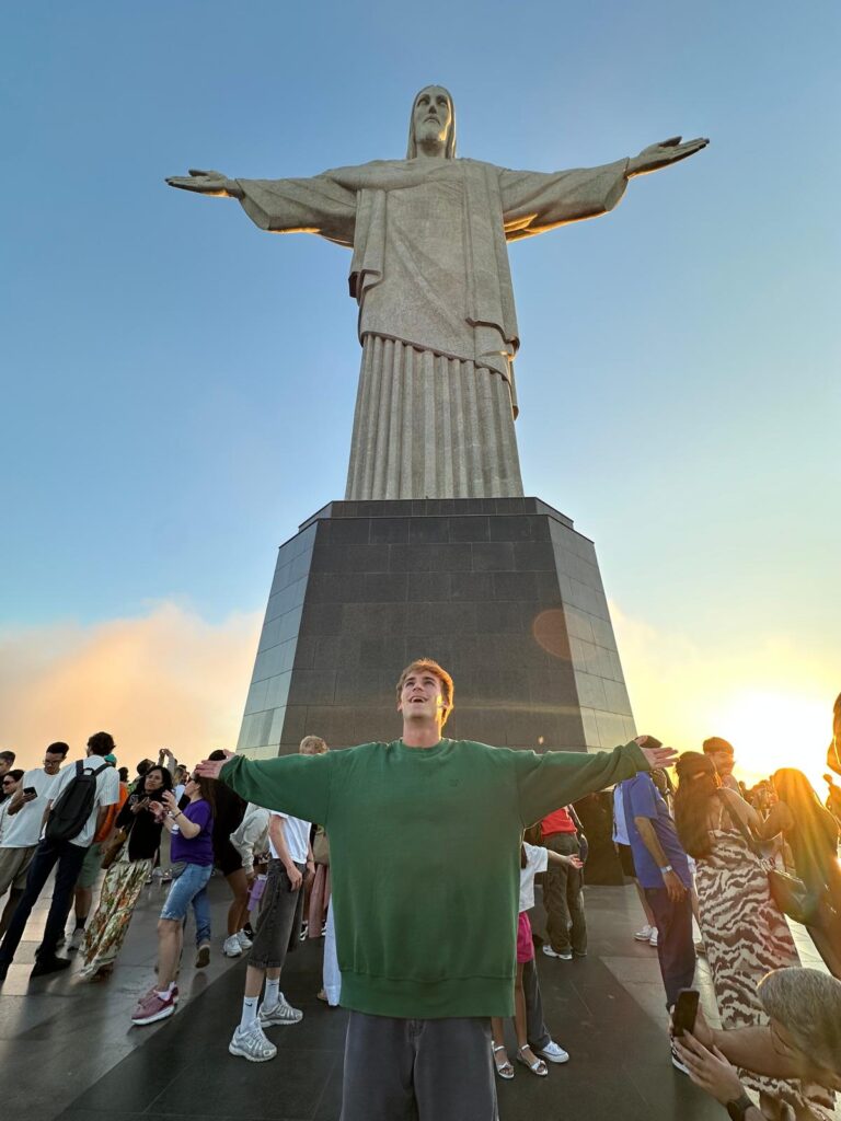 Alex stands in front of the "Christ the Redeemer" statue in Rio de Janeiro with him arms spread, mimicking the statue's pose.