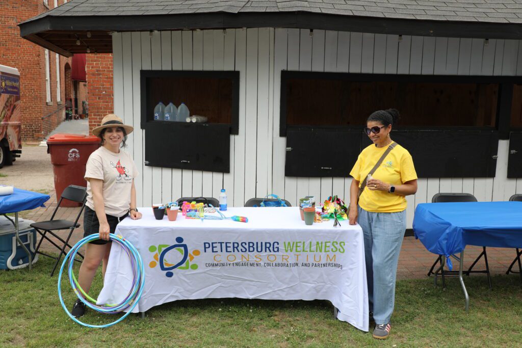 Image of 2 people standing near a table with a tablecloth reading "Petersburg Wellness Consortium"