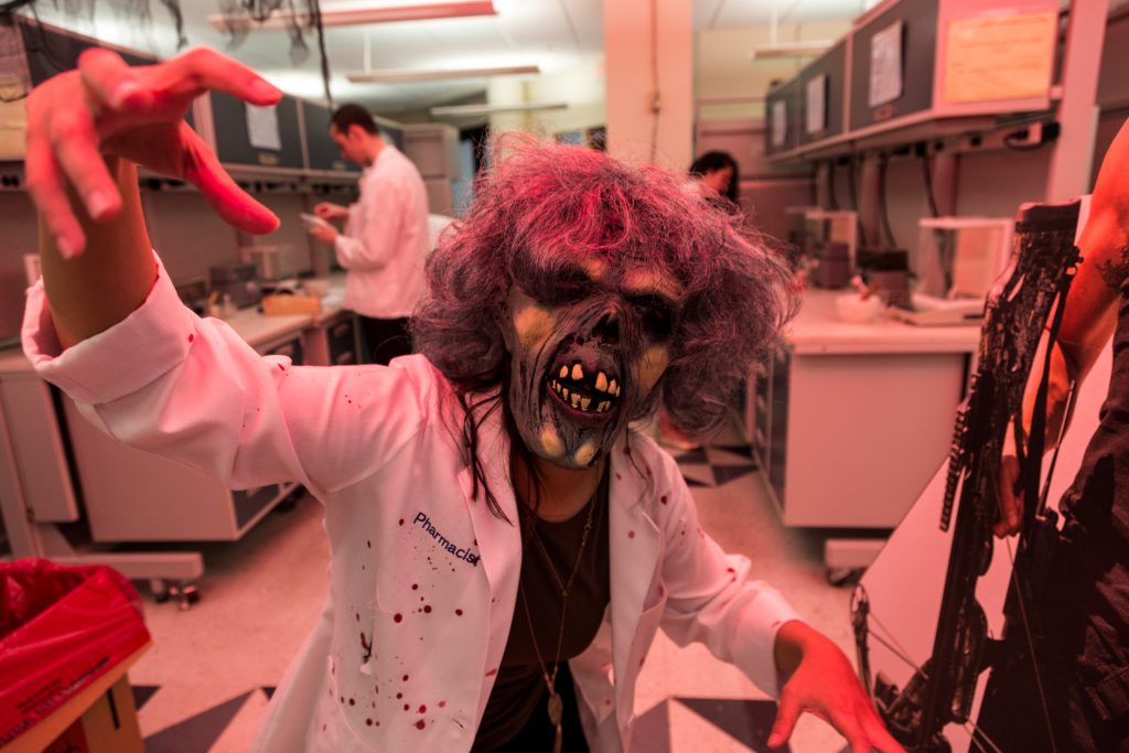 A fourth-year pharmacy student in a zombie mask and white coat spattered with fake blood pretends to threaten the camera.
