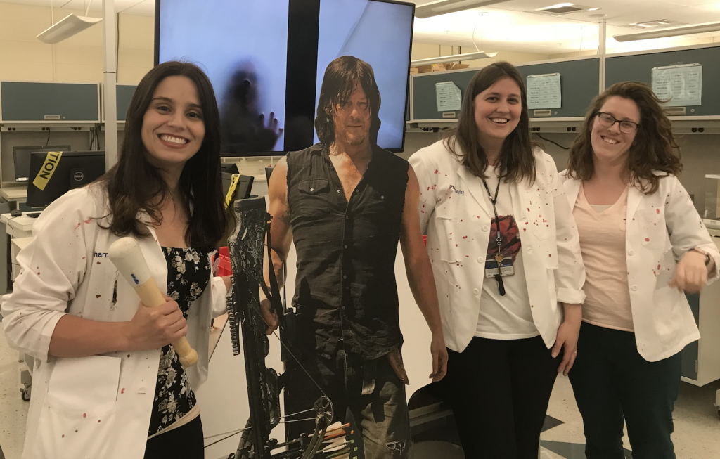 Three pharmacy professors -- Lauren Caldas, Krista Donohoe, Abigale Matulewicz -- pose with a cutout of "Walking Dead" actor Norman Reedus. All three professors wear white lab coats; a large LED screen behind them shows a creepy silhouetted hand. 