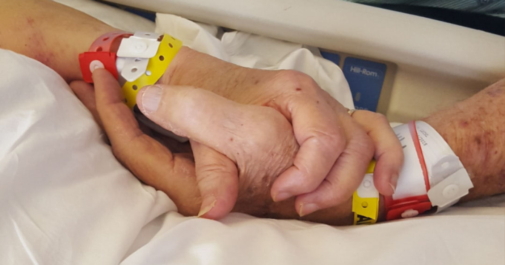 A photo of an old person's hand with a hospital wrist band being held by a younger person's hand.