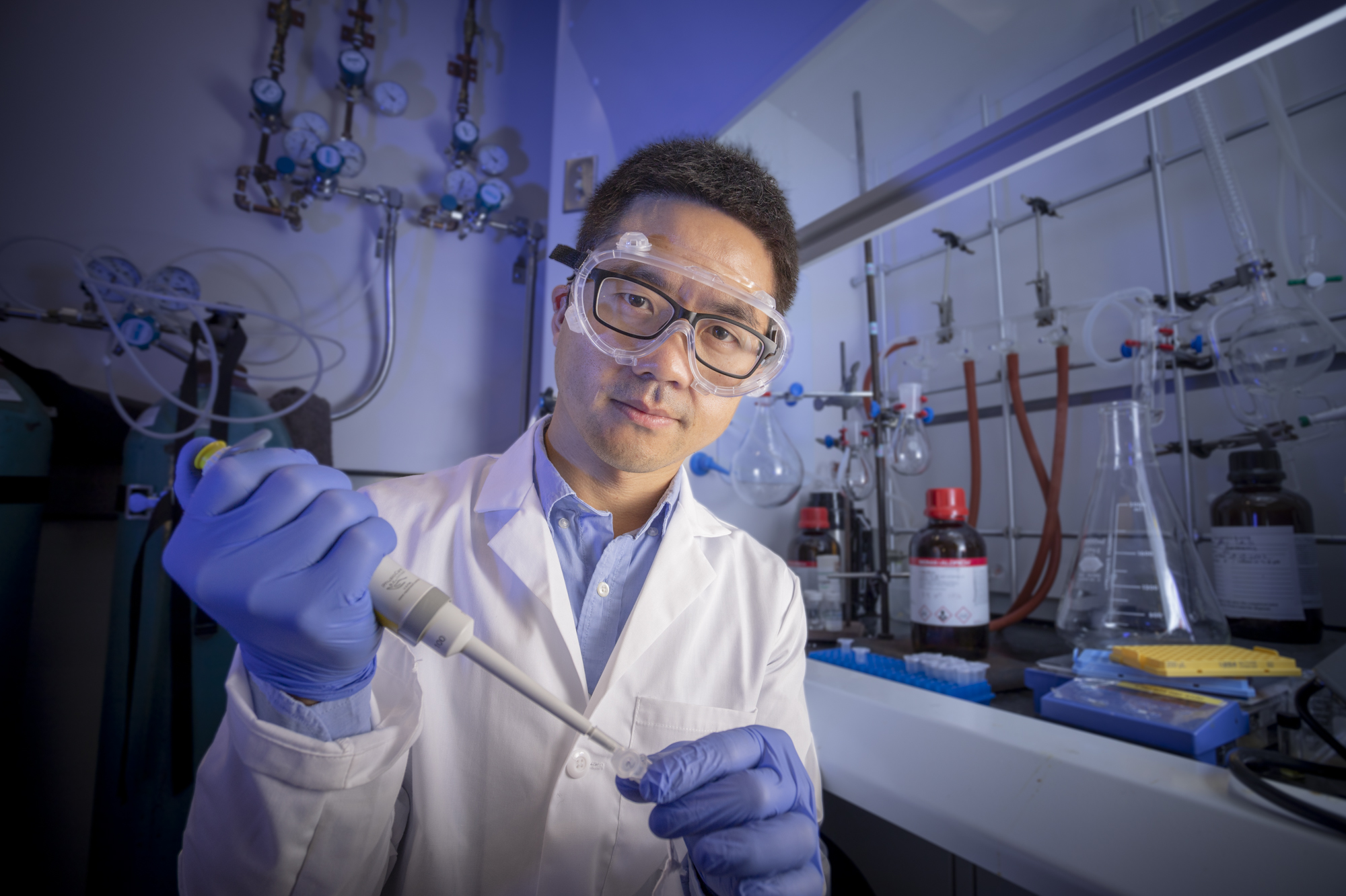 Julian Zhu in a white lab coat and blue gloves holds a pipette.