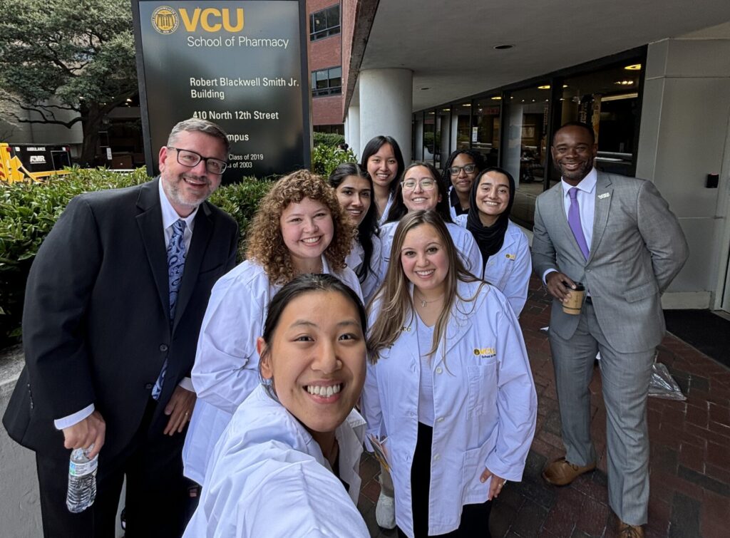 A group of students pose for a selfie with two men in suits, the dean of VCU School of Pharmacy and the APhA CEO.