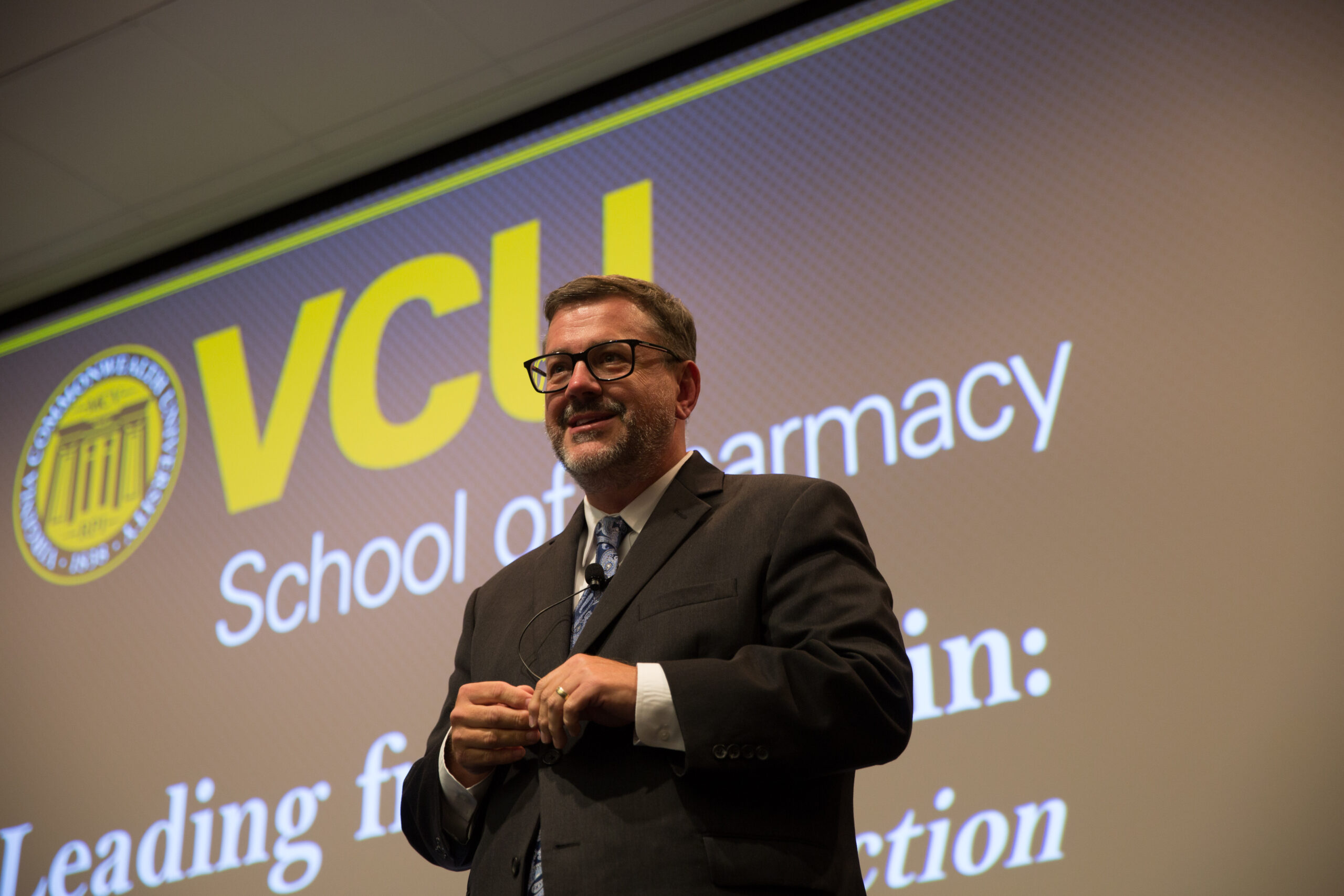 A man in a suit presents to a crowd while standing in front of a screen that says "VCU School of Pharmacy. Leading from Within: Agency, Advocacy and Action."