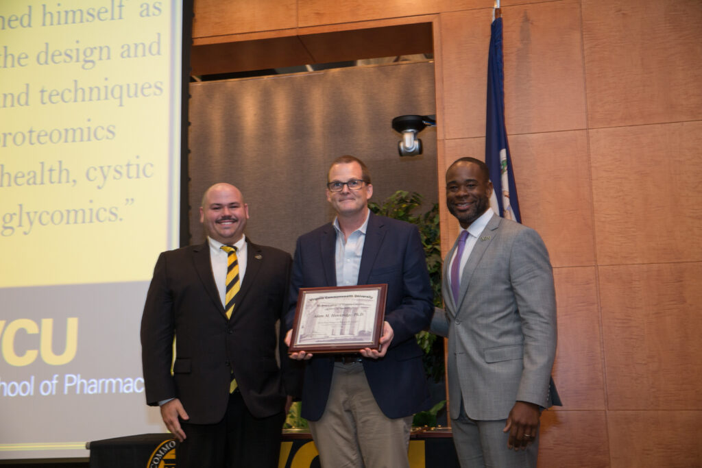 Adam Hawkridge, a man in a suit holding an award, smiles for a photo flanked by two smiling men in suits