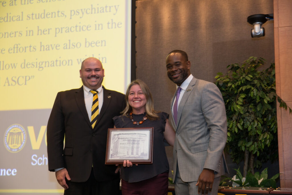 Ericka Crouse, a woman in a dress holding an award, smiles for a photo flanked by two smiling men in suits