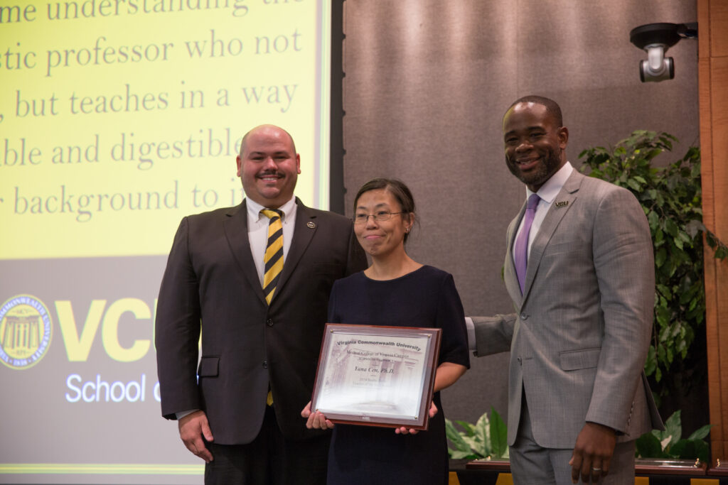 Yana Cen, a woman in a dress holding an award, smiles for a photo flanked by two smiling men in suits