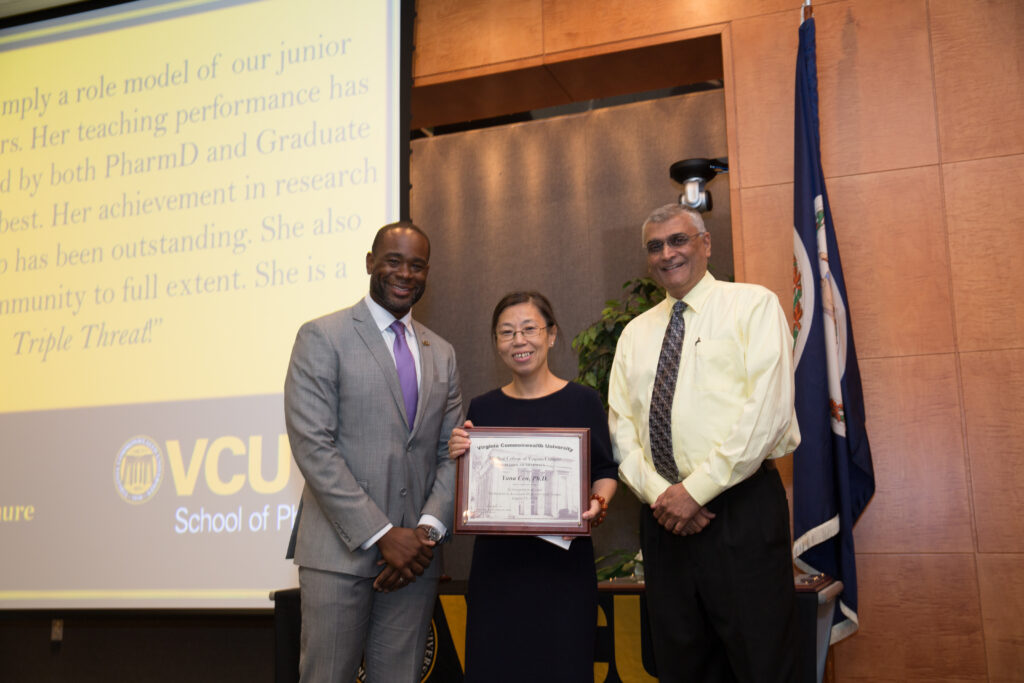 Yana Cen, a woman in a dress holding an award, smiles for a photo flanked by two smiling men in suits