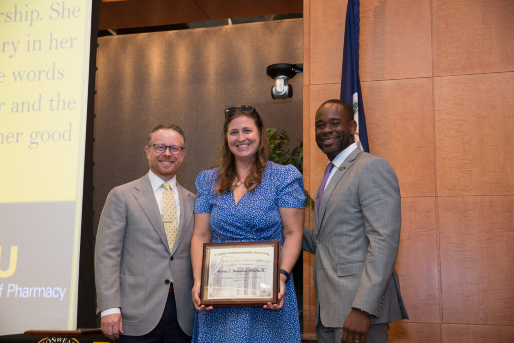 Krista Donohoe, a woman in a dress holding an award, smiles for a photo flanked by two smiling men in suits