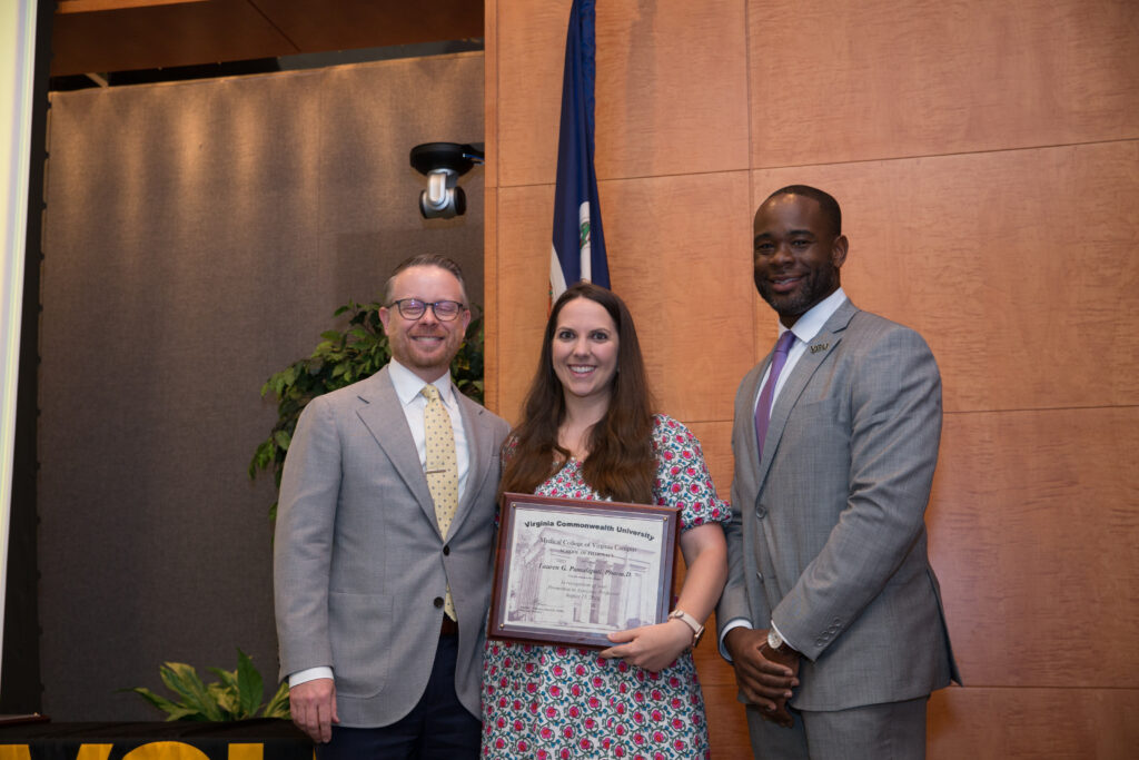 Lauren Pamulapati, a woman in a dress holding an award, smiles for a photo flanked by two smiling men in suits