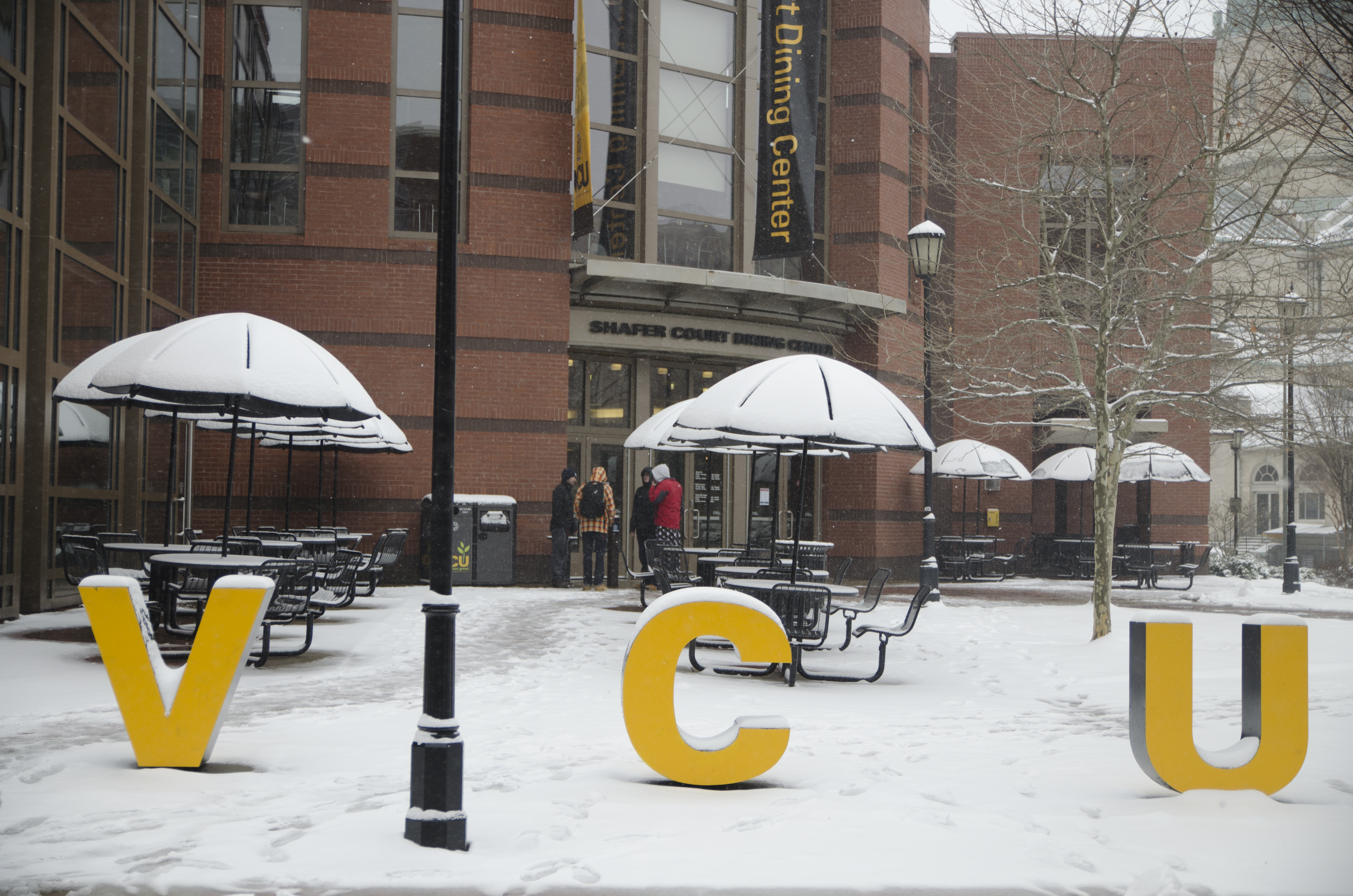 Outside Shafer Court on a snowy day. The gold VCU letters have snow on top of them. Four students are standing in the entryway of Shafer Court Dining Center.