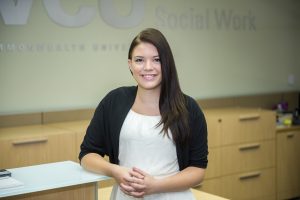Carli M. Bostic stands in front of a desk with the VCU Social Work logo behind her